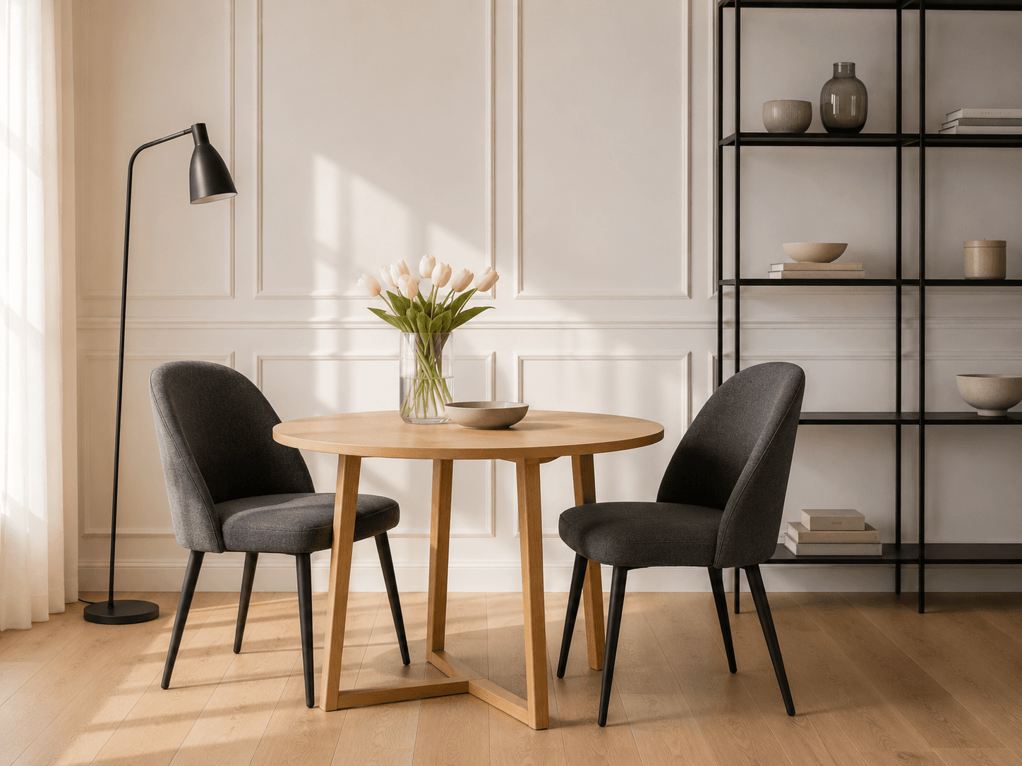Round wooden dining table with two dark upholstered chairs, a floor lamp and a shelving unit